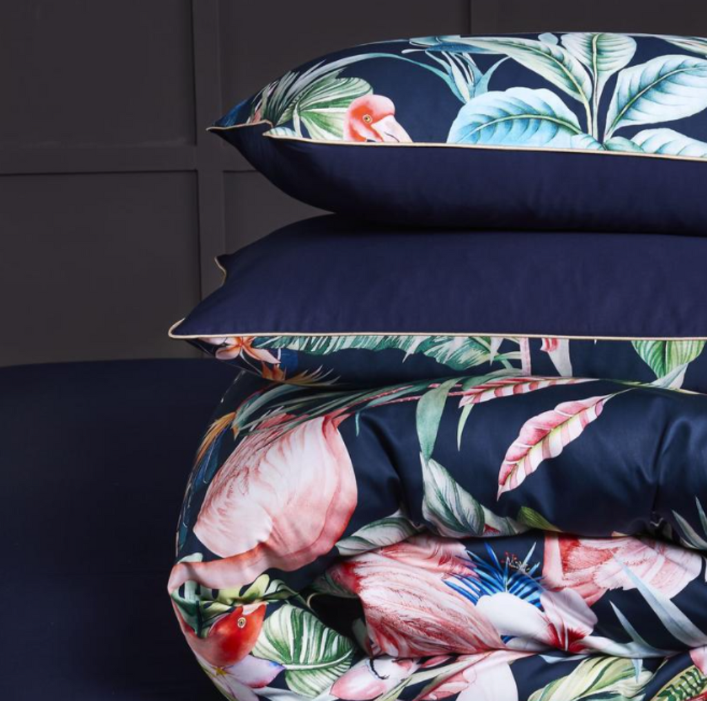 Two botanical flamingo pillows resting on a folded tropical duvet atop navy flat sheet — showcasing layered texture and vibrant print.